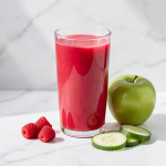 Glass of pink raspberry cucumber apple juice with fresh raspberries, cucumber slices and a green apple on a white marble surface