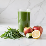 Glass of fresh green arugula apple and cucumber juice on a marble counter with rocket leaves, two red apples, half a lemon and a cucumber beside it