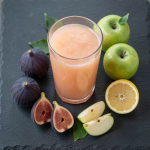 Fresh fig and apple juice in a glass with ripe purple figs, green apples and a halved lemon on a dark slate board