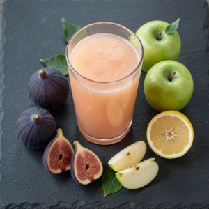 Fresh fig and apple juice in a glass with ripe purple figs, green apples and a halved lemon on a dark slate board