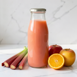 A sealed bottle of freshly pressed rhubarb, apple and orange juice on a white marble surface with three whole rhubarb stalks, a red apple, a golden apple and a halved orange beside it