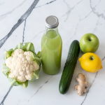 Cauliflower, cucumber, apple and lemon juice in a sealed glass bottle on a white marble surface, with a whole white cauliflower head, a cucumber, green apples, a lemon and ginger root beside it